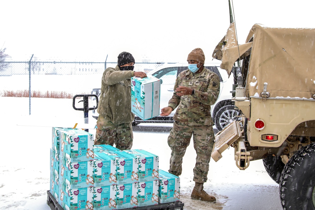 A soldier passes a box of diapers to another soldier standing by the back of a military vehicle in a parking area.
