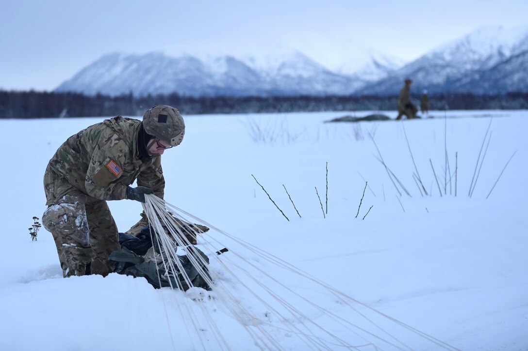 A soldier gathers a parachute in a snow-covered field with mountains in the background.