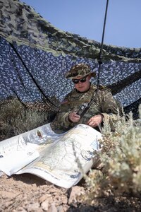 A soldier sits under coma netting and radios coordinates from a map