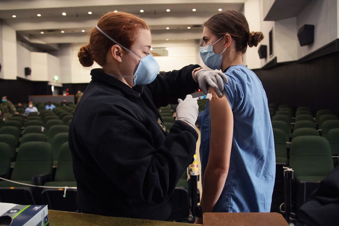 A soldier wearing a face mask and gloves administers a COVID-19 vaccine to a Navy petty officer wearing a face mask.
