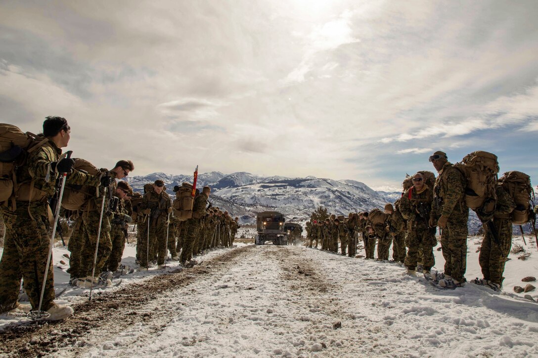 Marines stand in line and wait for a vehicle in the snow.