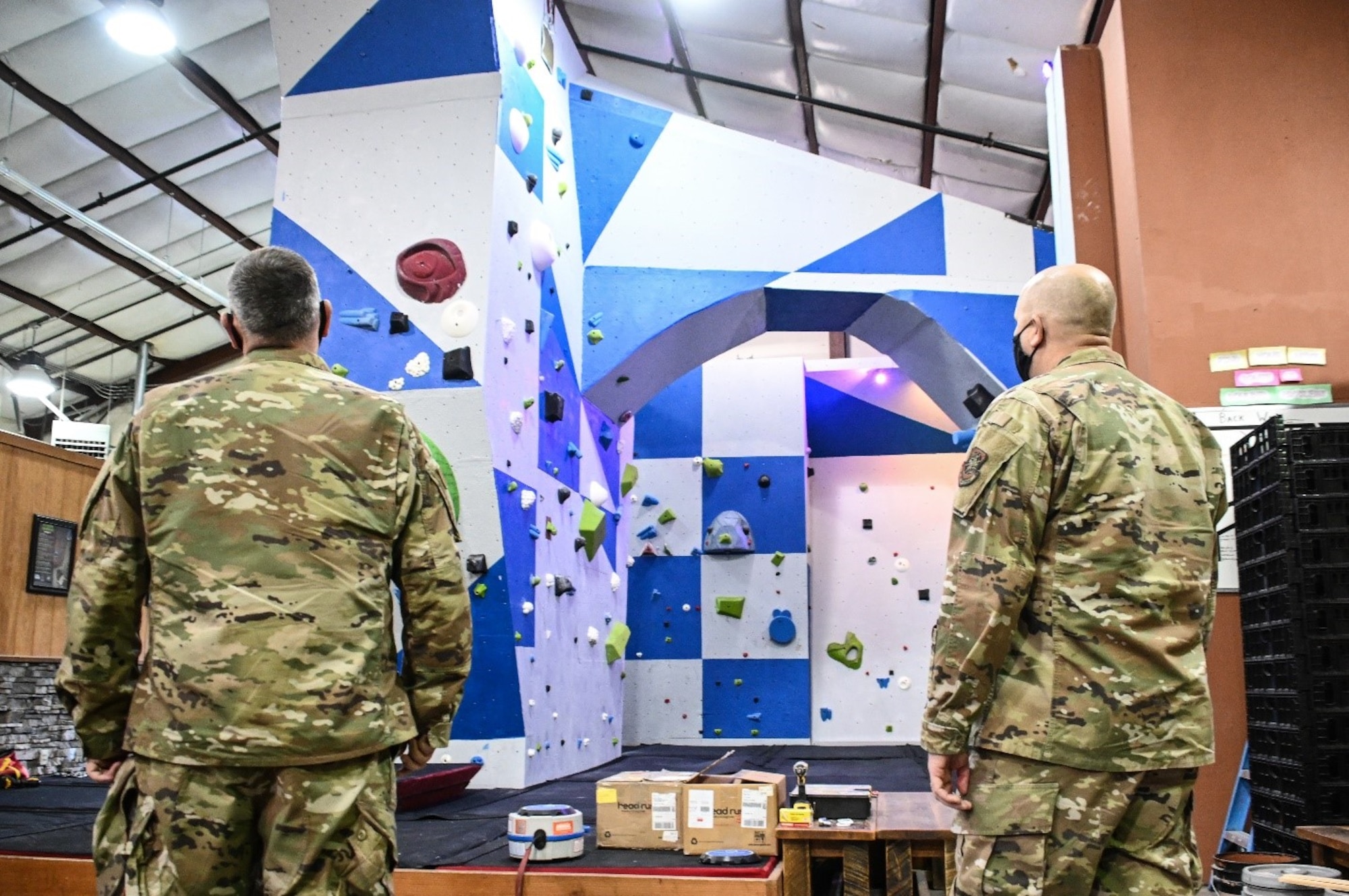 Two men in military uniforms look up at an indoor rock climbing wall