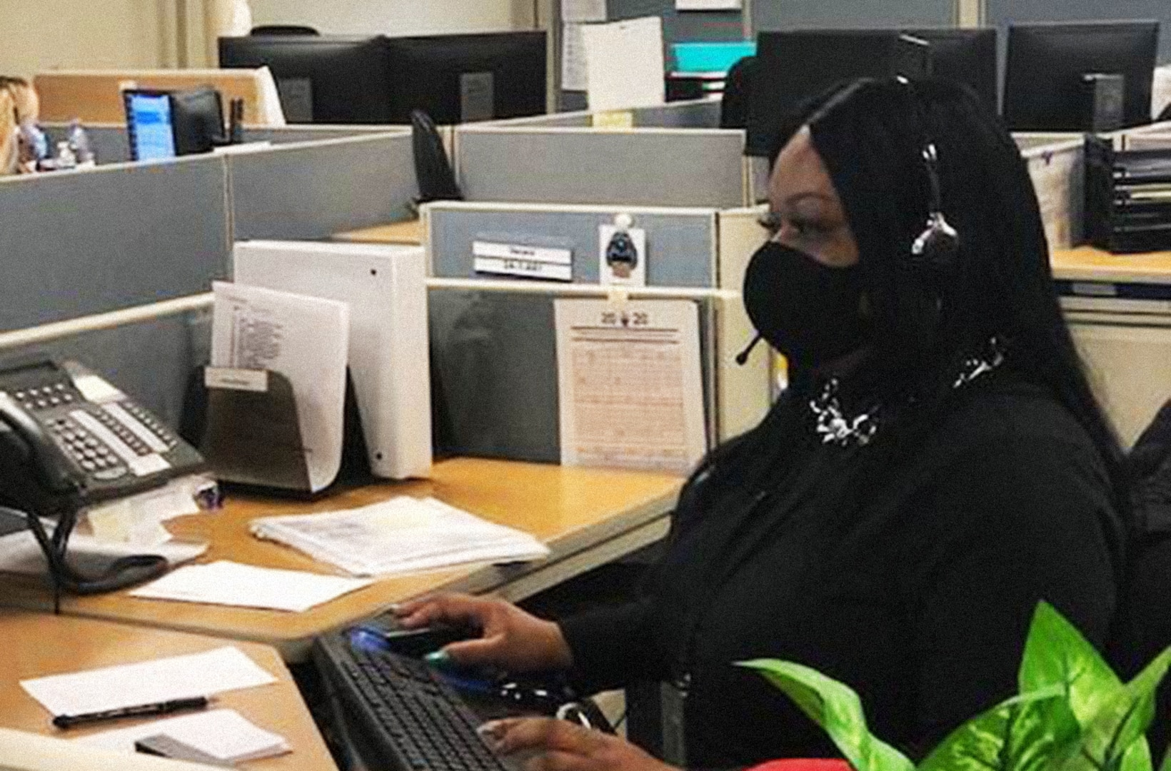 Black woman wearing a black shirt, face mask and headset sits at a desk in front of a computer.