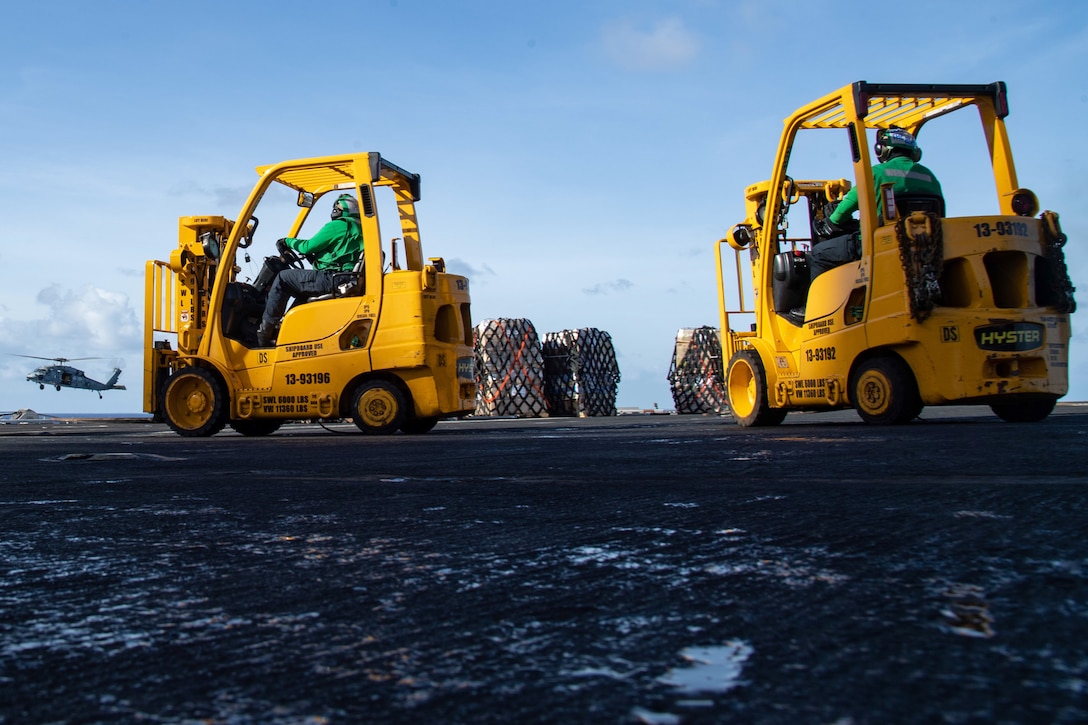 Sailors drive two yellow forklifts on a ship's flight deck as a helicopter flies low in the background.
