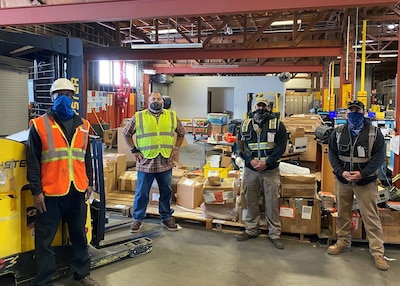 Four men wearing personal protective equipment for warehouse work and facemask stand socially distanced inside a warehouse with a forklift and boxes on pallets around them.
