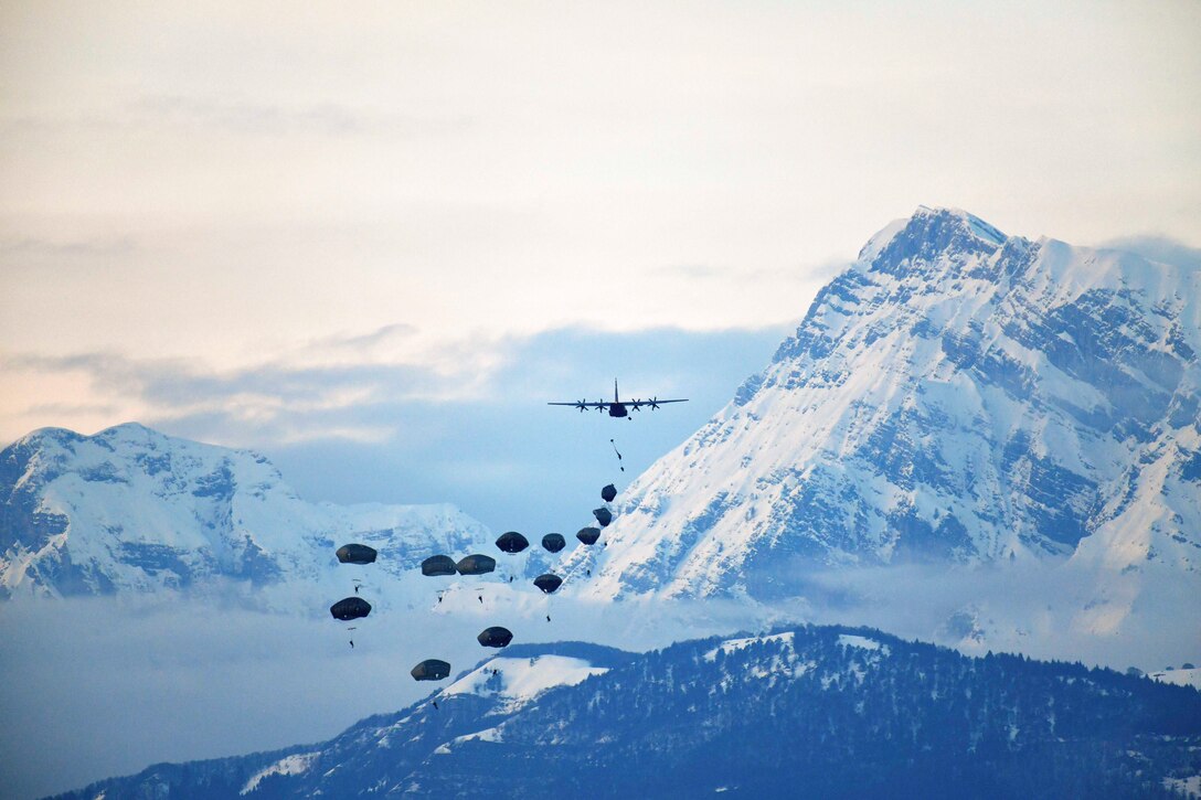 Soldiers freefall with parachutes after exiting an aircraft near mountains.