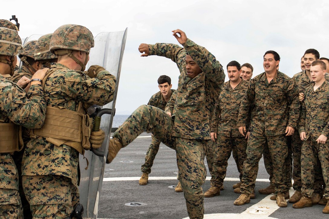 A Marine kicks a shield held by a fellow Marine as others watch on a ship's deck.