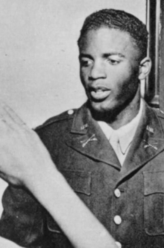 A young man is dressed in a military uniform; the arm of someone saluting is seen in the foreground.