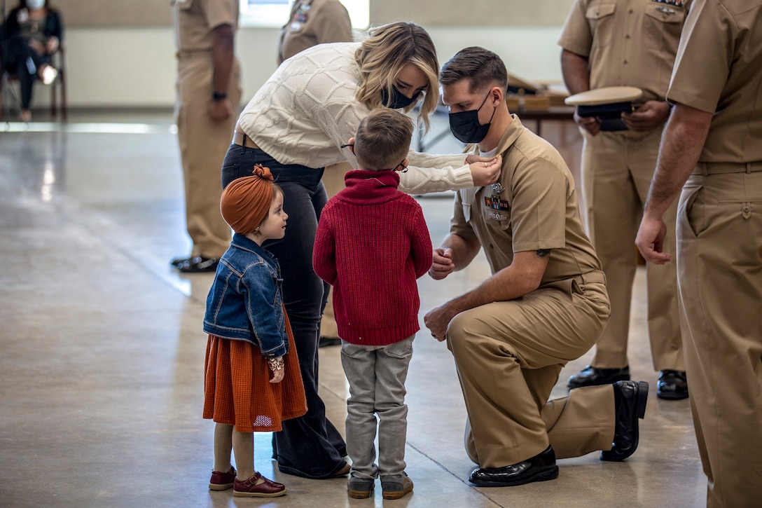 A sailor kneels as a civilian puts a pin on his uniform collar as two children watch.