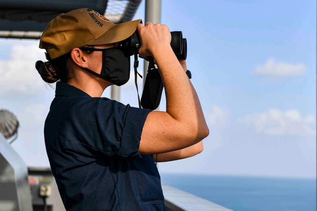 A sailor looks through binoculars while standing on a ship.