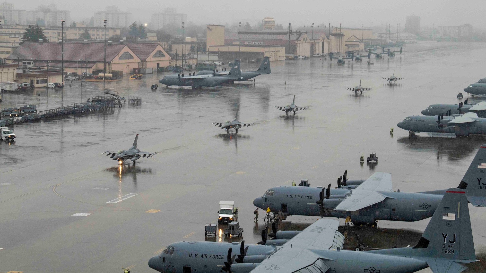 A wide shot with C-130's in the foreground and a line of F-16s taxing down a rainy flightline.