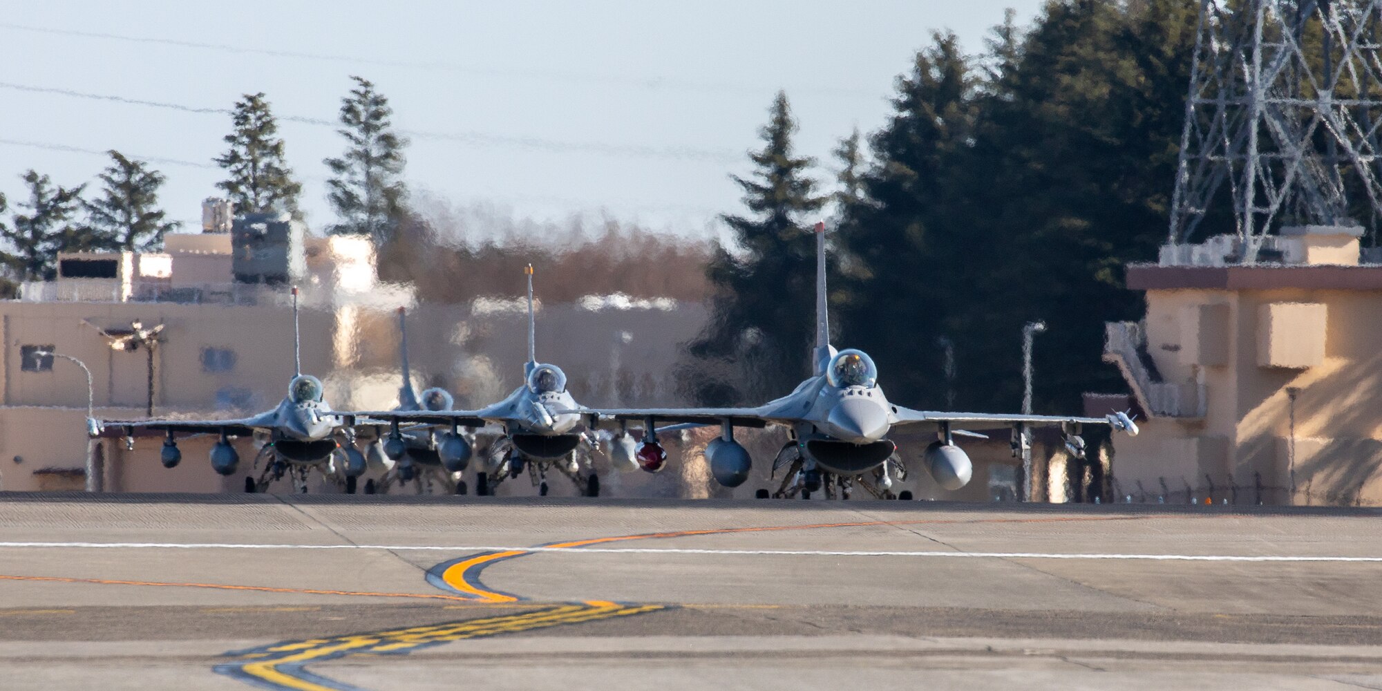 Three F-16s taxi up the flightline in a row. The heat from their engines creates blur in the air.