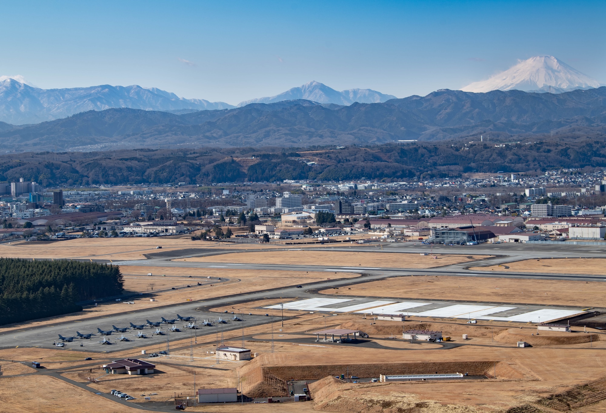 A wide photograph of the Yokota flightline with the town behind it and mountains beyond that. 15 F-16s sit on the flightline in the foreground.