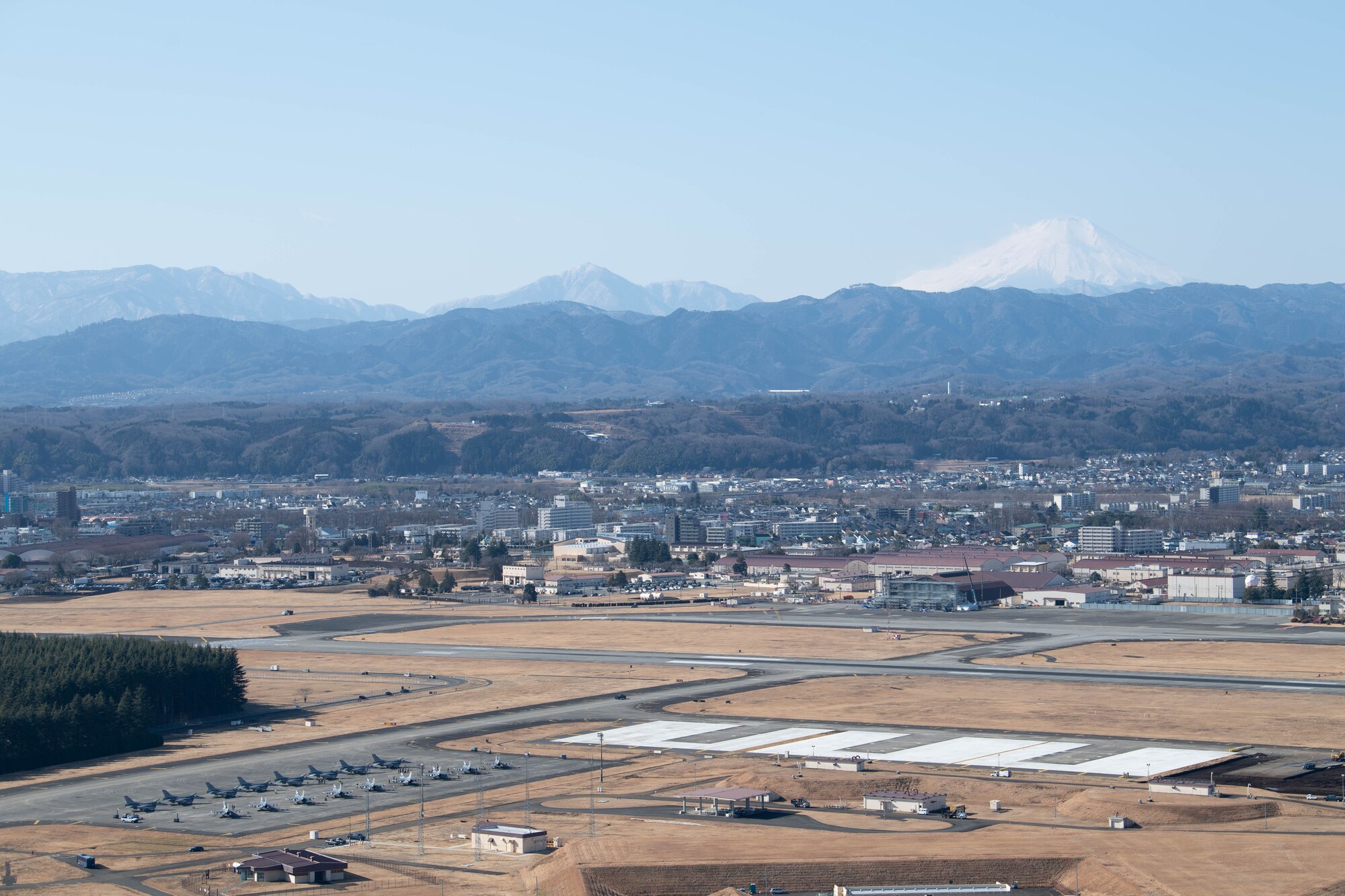 A wide photograph of the Yokota Air Base flightline, with the city behind it and the mountains beyond that. 15 F-16s sit on the flightline in the foreground.