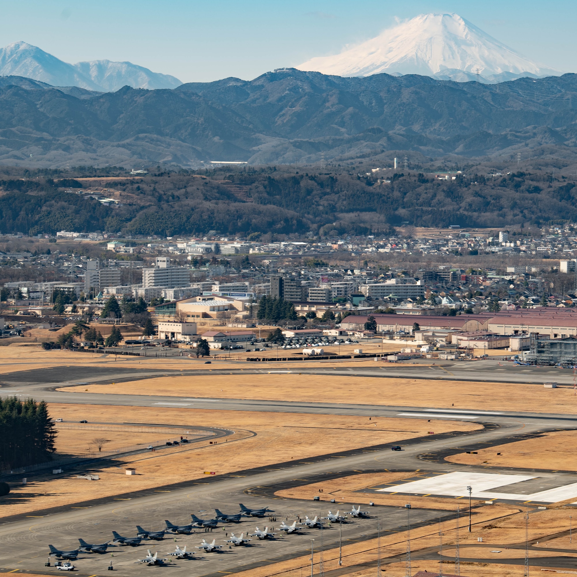Wide photo of the Yokota Flightline and the city behind it, with mountains beyond that. 15 F-16s sit on the flightline in the foreground.