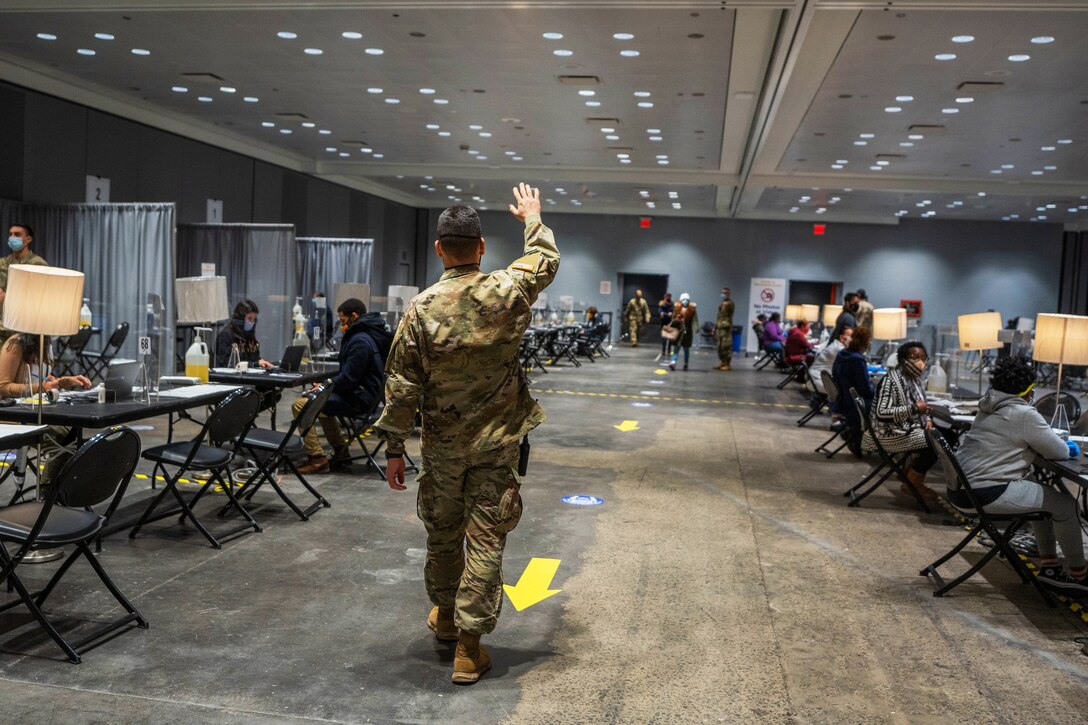 A soldier stands at the head of a series of arrows painted on a concrete floor; he motions to members of the public to walk toward him. People sit at long tables with chairs on each side of him.