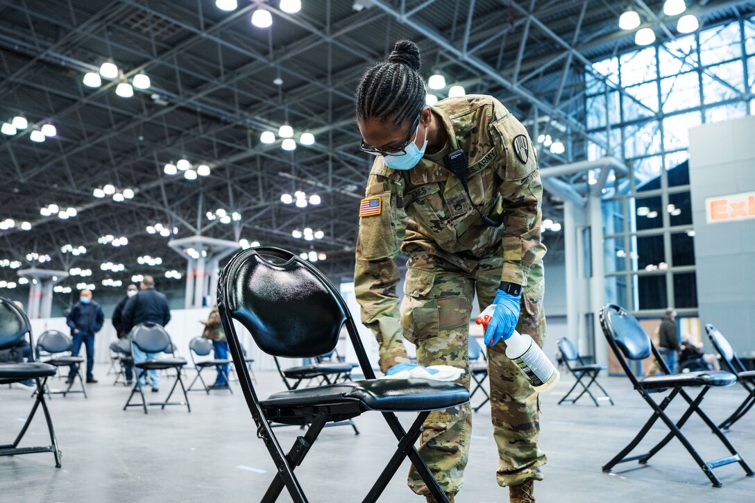 A soldier wearing a face mask and gloves wipes the seat of a chair with a cloth. In her other hand, she holds a spray bottle.