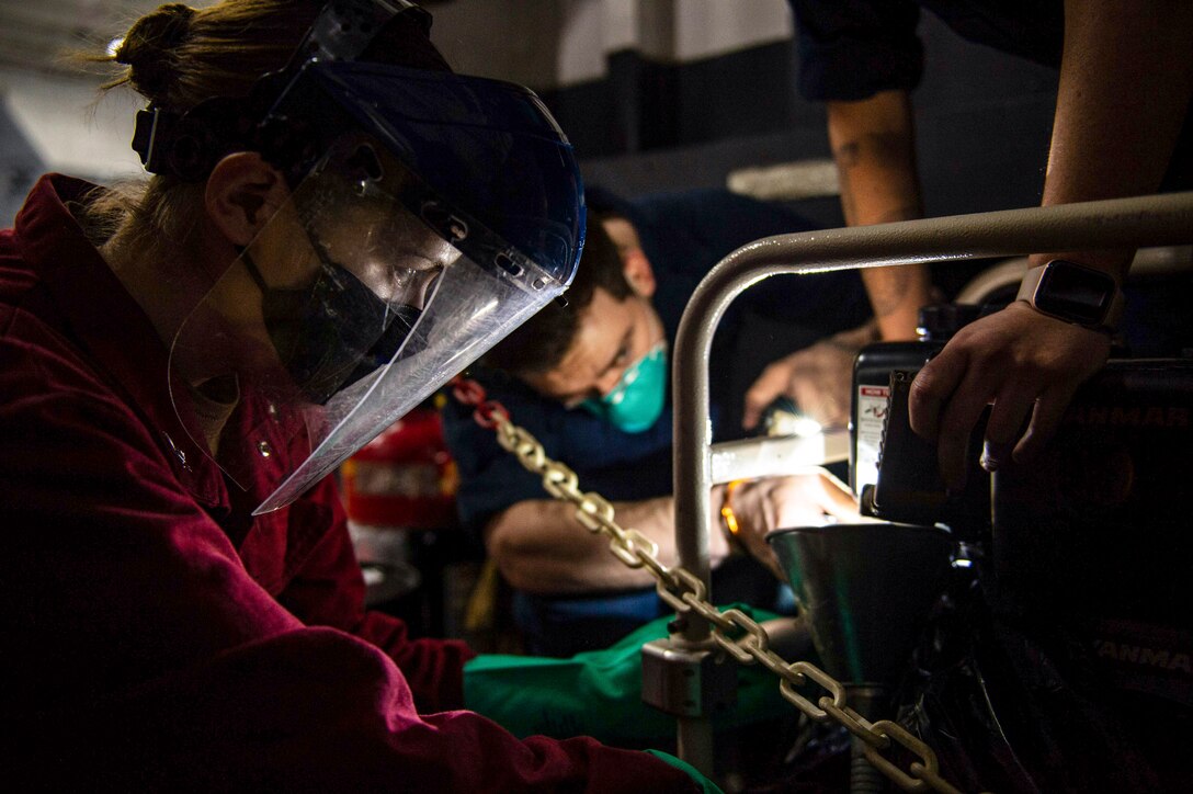 Three sailors work on a piece of equipment in a dark area.