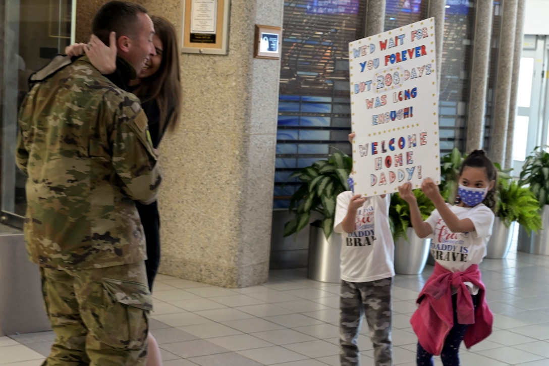 An airmen hugs his wife while looking at poster board his children are holding.