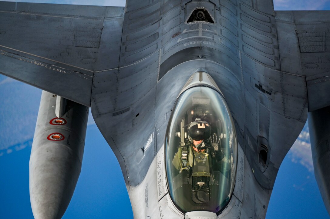 An Air Force pilot, seen from above, signals from the cockpit of a jet flying in blue skies.