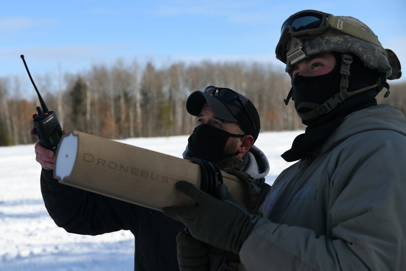 Two men stand next to each other as one holds a anti-drone device.