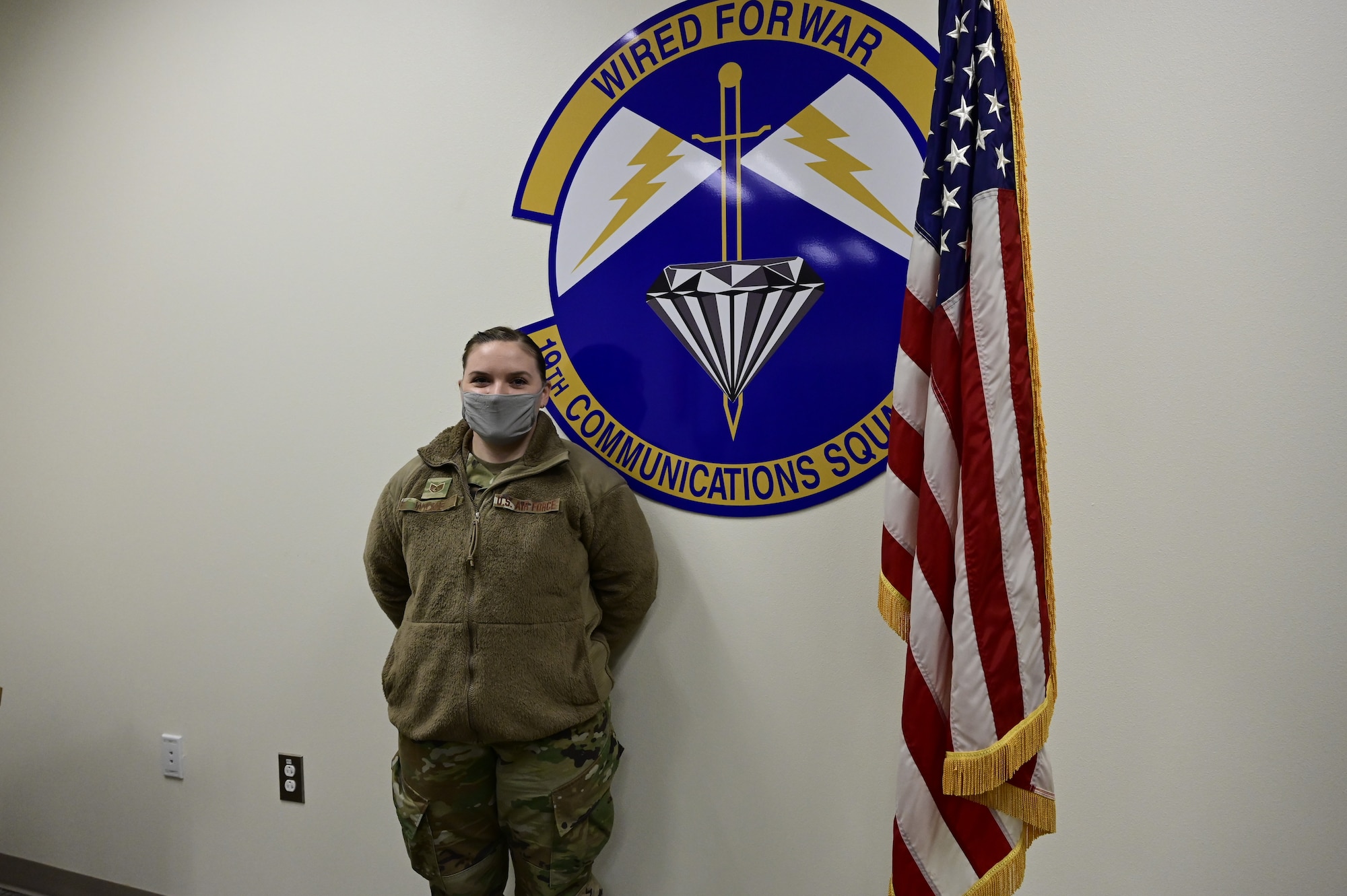 An Airman poses for a photo in front of a 19th Communications Squadron badge