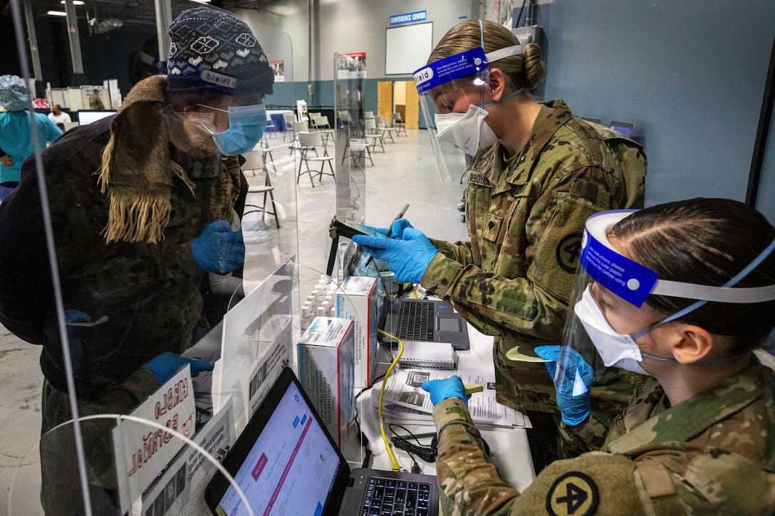 Two soldiers wearing face masks and gloves review information with a person on the other side of a glass shield wearing a face mask and gloves.