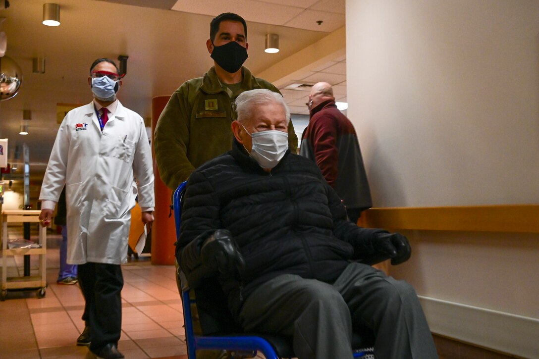 An Air Force officer wearing a face mask pushes an elderly man in a wheelchair wearing a face mask down the hall to a vaccine clinic.