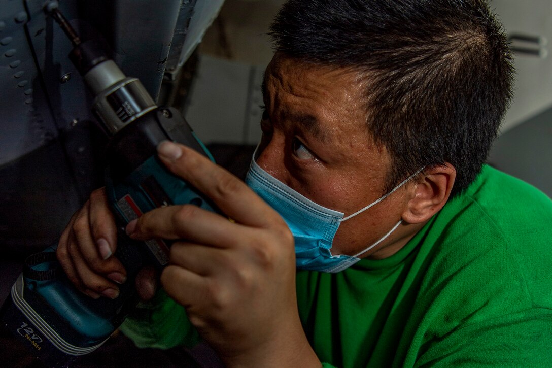 A sailor in a green shirt uses a drill to tighten a screw on the body of an aircraft.