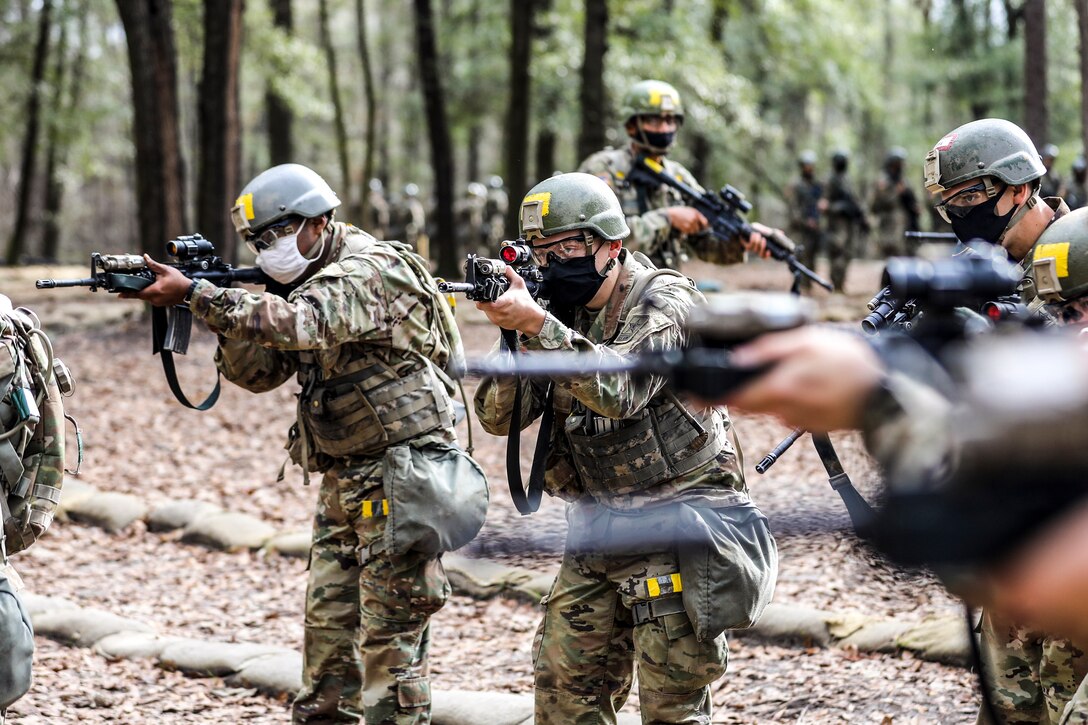 Soldiers aim weapons in a forested area.