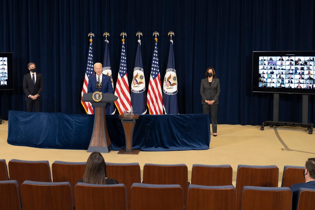 A man stands on a stage at a lectern with microphones with two people socially distanced on either side of him. Four flags are in the background and the audience is socially distanced.