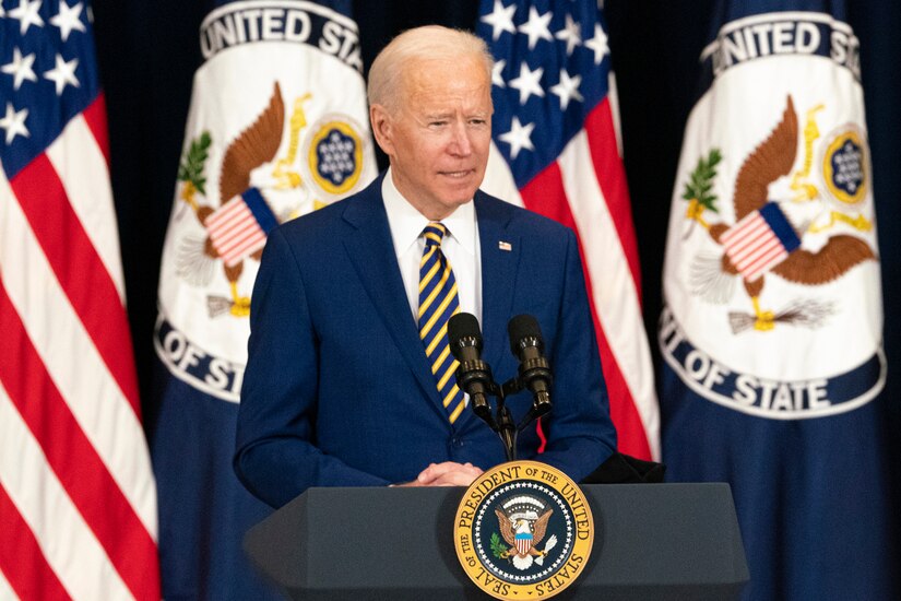 A man stands at a lectern with microphones and the Seal of the President of the United States affixed to the lectern. Four flags are in the background.