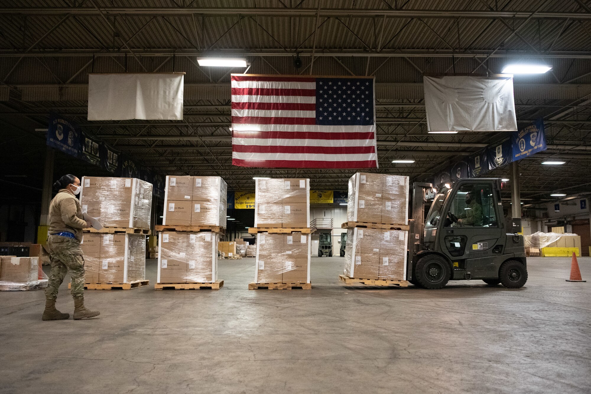 A fork lift is moving wood pallets from one place to another under a flag inside a warehouse.