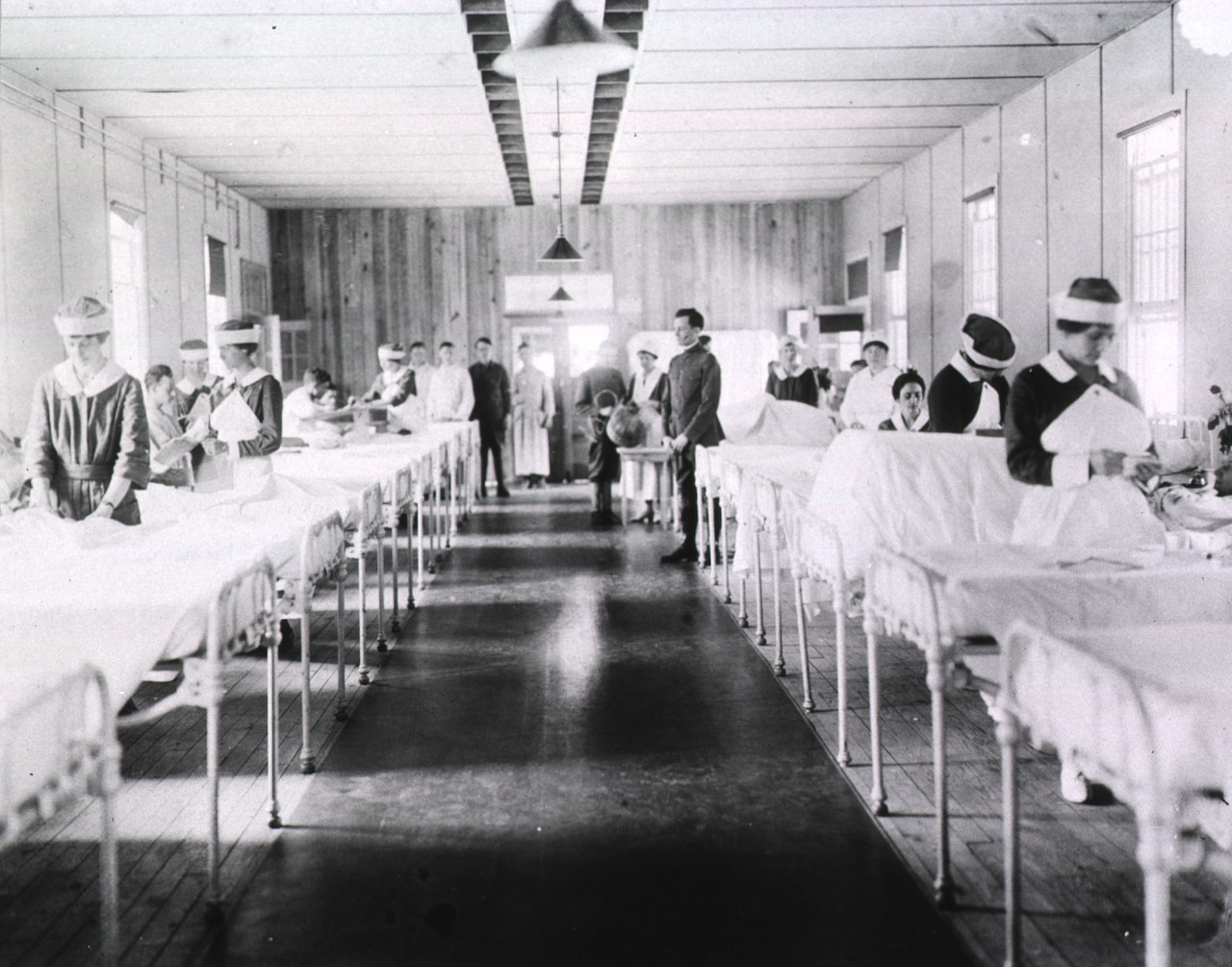 Several hospital beds are lined up in a room. Nurses tend to some of the patients in those beds.