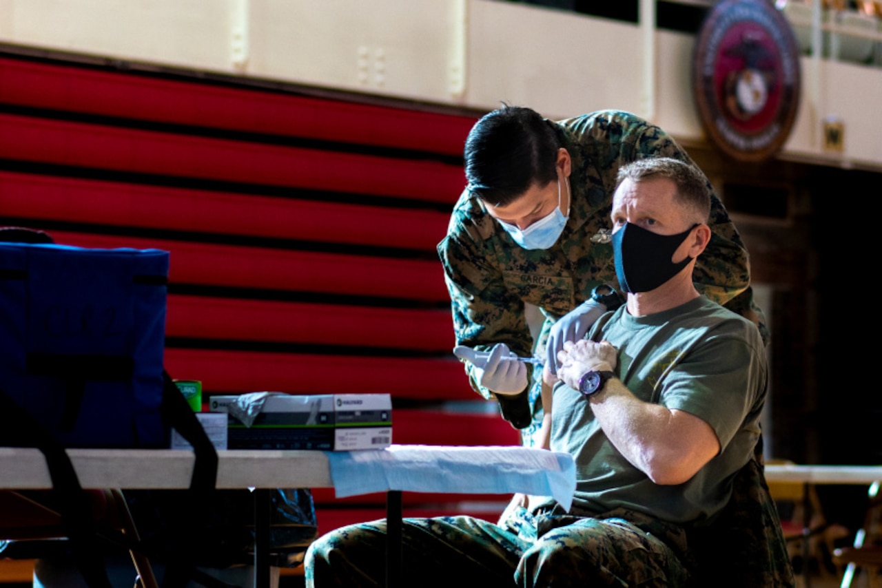 An older service member sits in a chair while a younger service member  leans down and administers the vaccine.