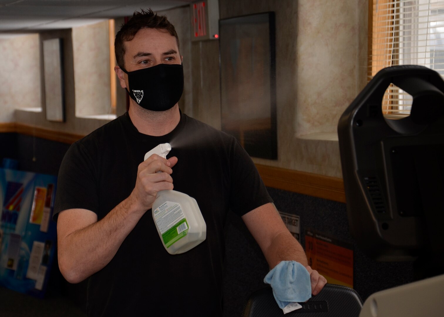 A person with a mask covering his mouth and nose sprays cleaner at fitness equipment to sanitize it for the next person.