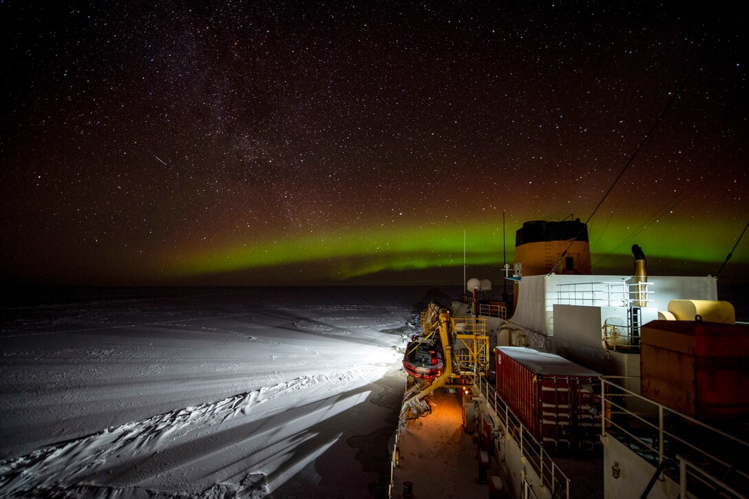 A Coast Guard icebreaker breaks travels through icy water in a night sky illuminated by an aurora.