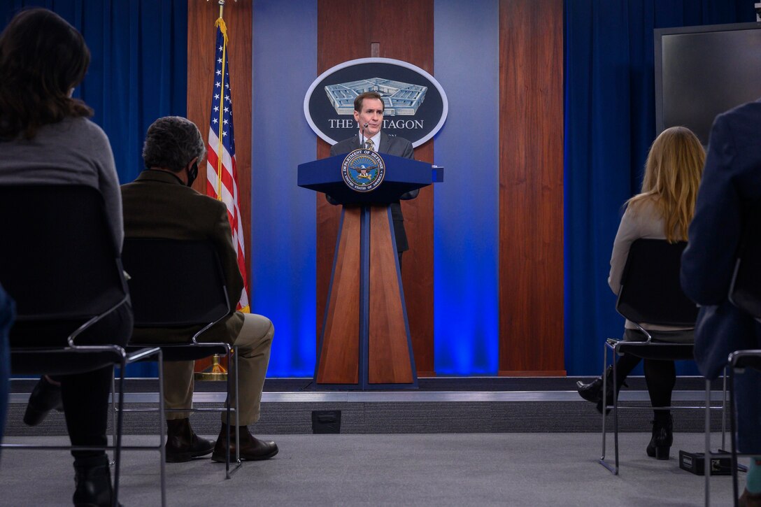 A man standing at a lectern briefs members of the press.