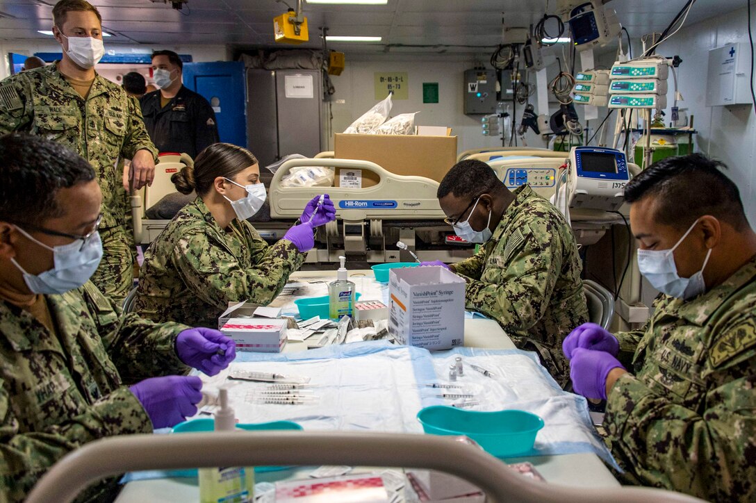 Sailors in masks and purple surgical gloves sit at a table and prepare syringes.