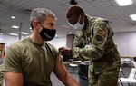 Female service member giving a COVID-19 vaccine to a male service member.