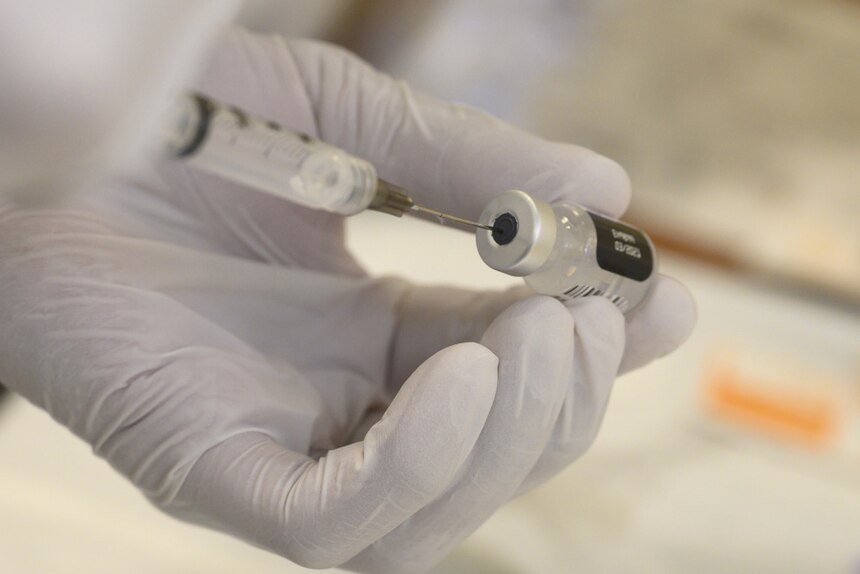 A gloved hand uses a syringe to draw fluid from a small bottle.