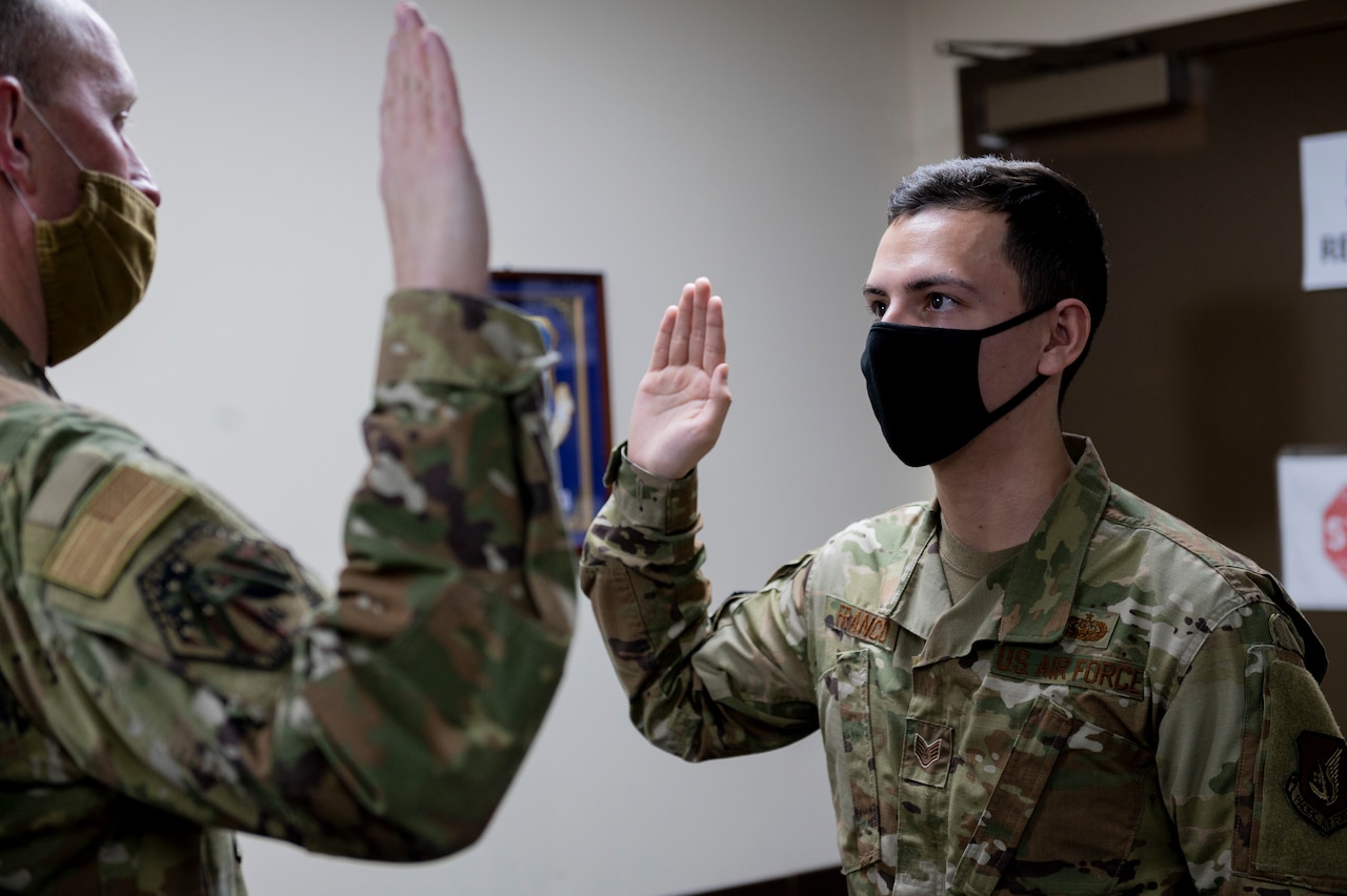 An airmen stands with his right hand raised facing another airman doing the same.