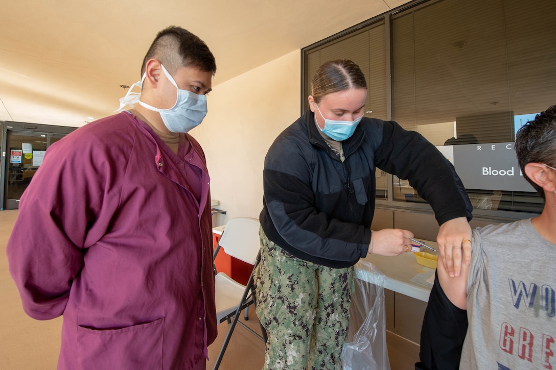 A sailor wearing a face mask gives another sailor on the job training administering a COVID-19 vaccine.