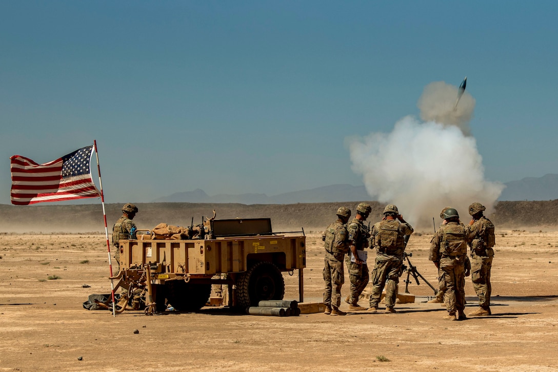 American and French soldiers fire a weapon while standing next to a trailer flying the  American flag.