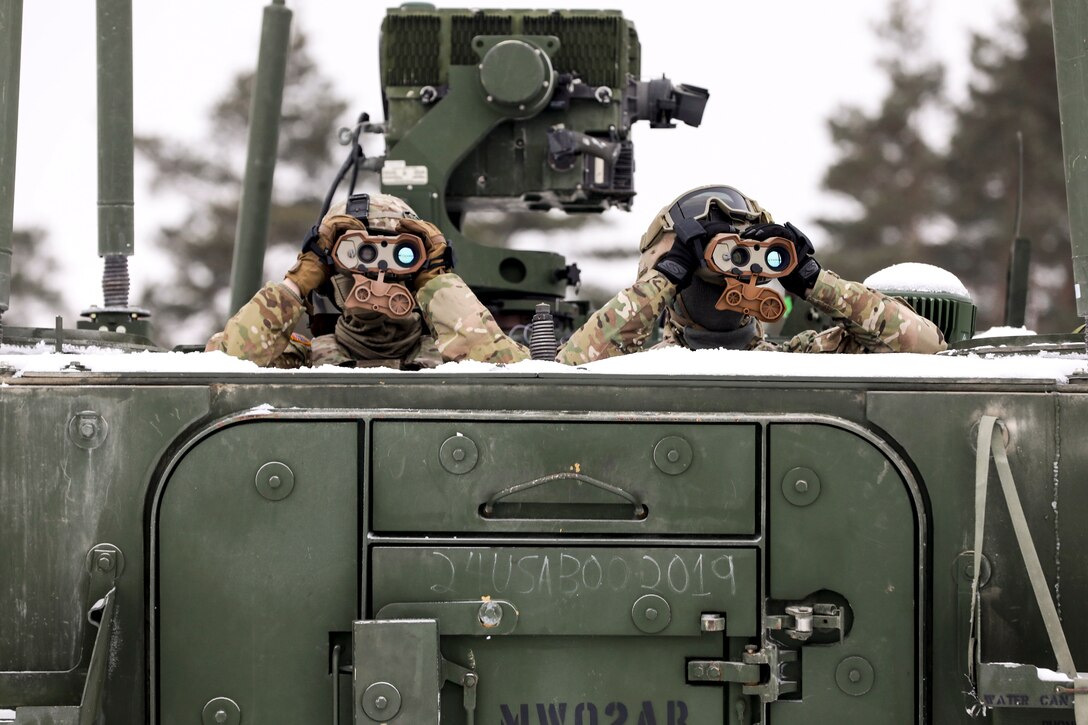 Two soldiers look through binoculars while covered up to their shoulders by a piece of equipment.