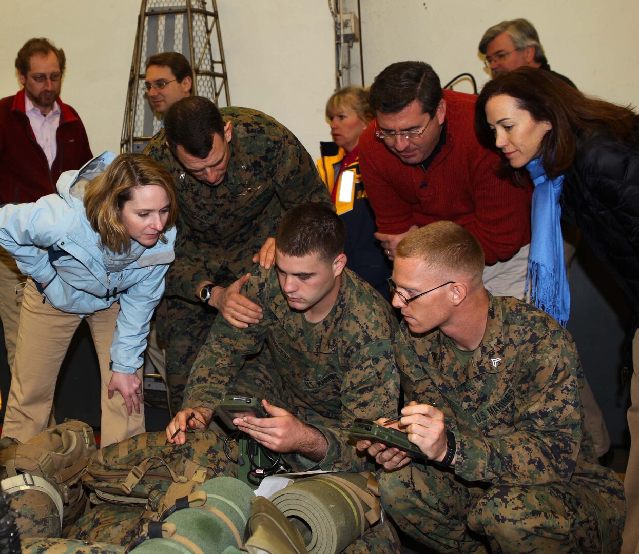 Three civilians lean over to look at a small device being held in the hand of a service member as other service members watch.