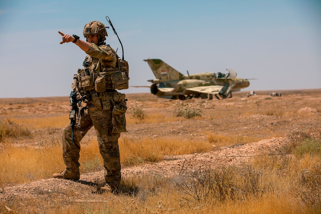 A soldier stands in a dry field and points at something in the distance; behind him, an old aircraft sits rusting in the desert.