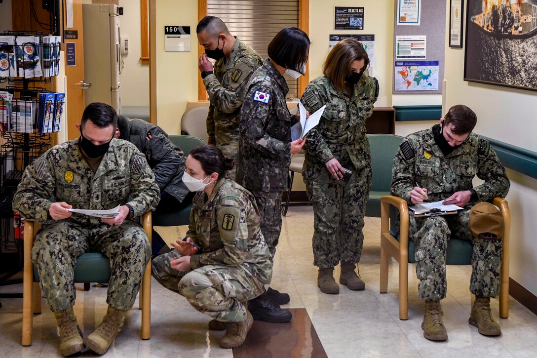 Three service members sit in a small room filling out paperwork. Three other service members lean over to watch; a fourth service member stands nearby holding papers.