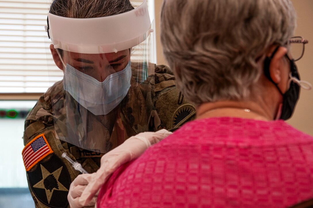 A soldier wearing a face mask, shield and gloves gives an elderly woman an injection.