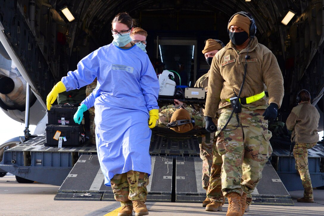 Four service members wearing face masks and gloves disembark from the cargo section of a large plane followed by two service members who are carrying someone on a stretcher.